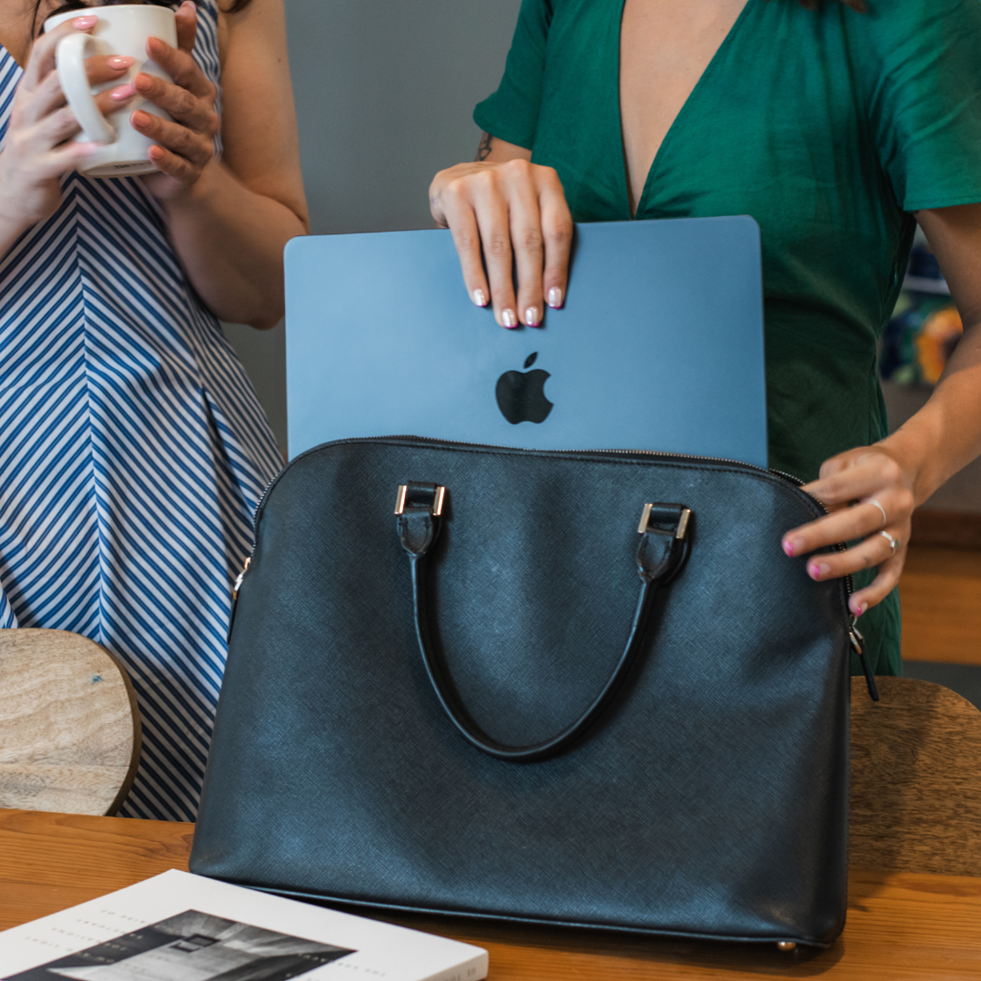 Two women using the Monday Concealed Carry Laptop Bag by Zendira—one holding coffee, the other sliding a MacBook into the structured black leather bag at a shared workspace.