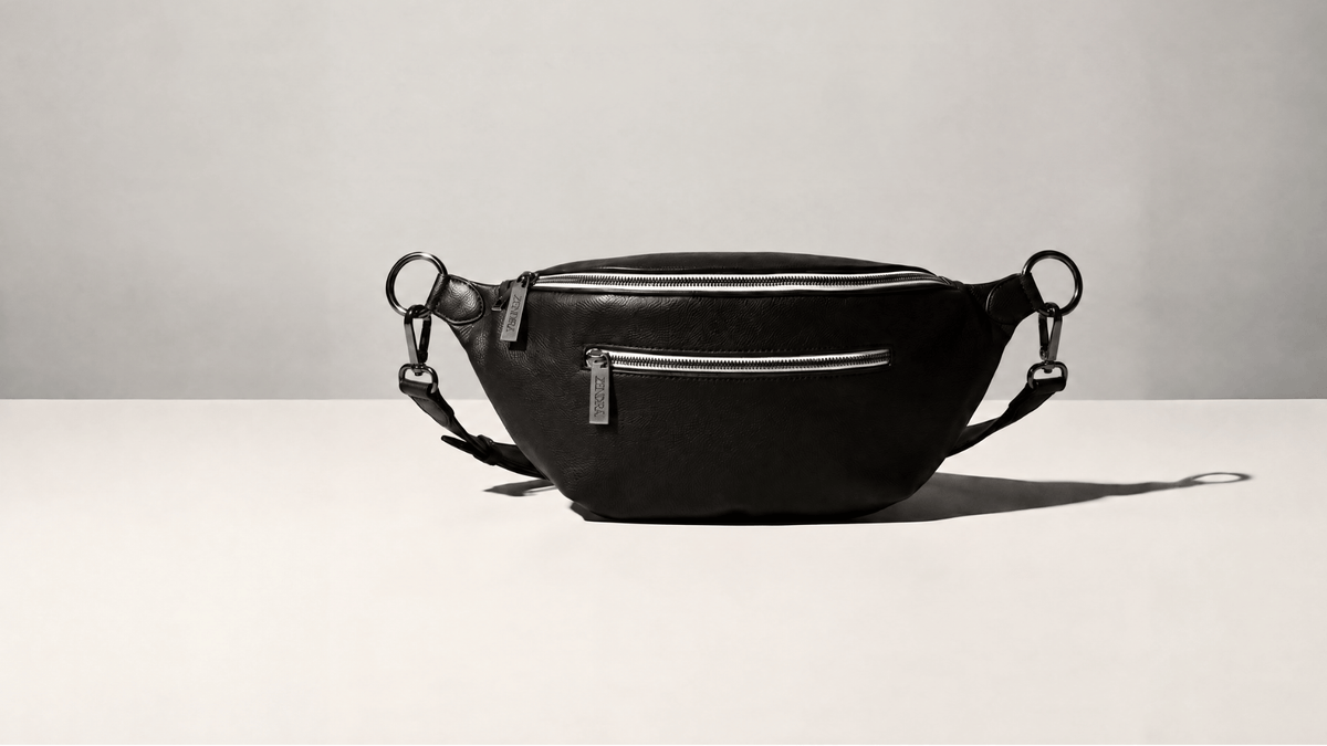 A black leather ZENDIRA bag with silver hardware, featuring a front zippered pocket and adjustable shoulder straps, displayed on a minimalist light gray background.