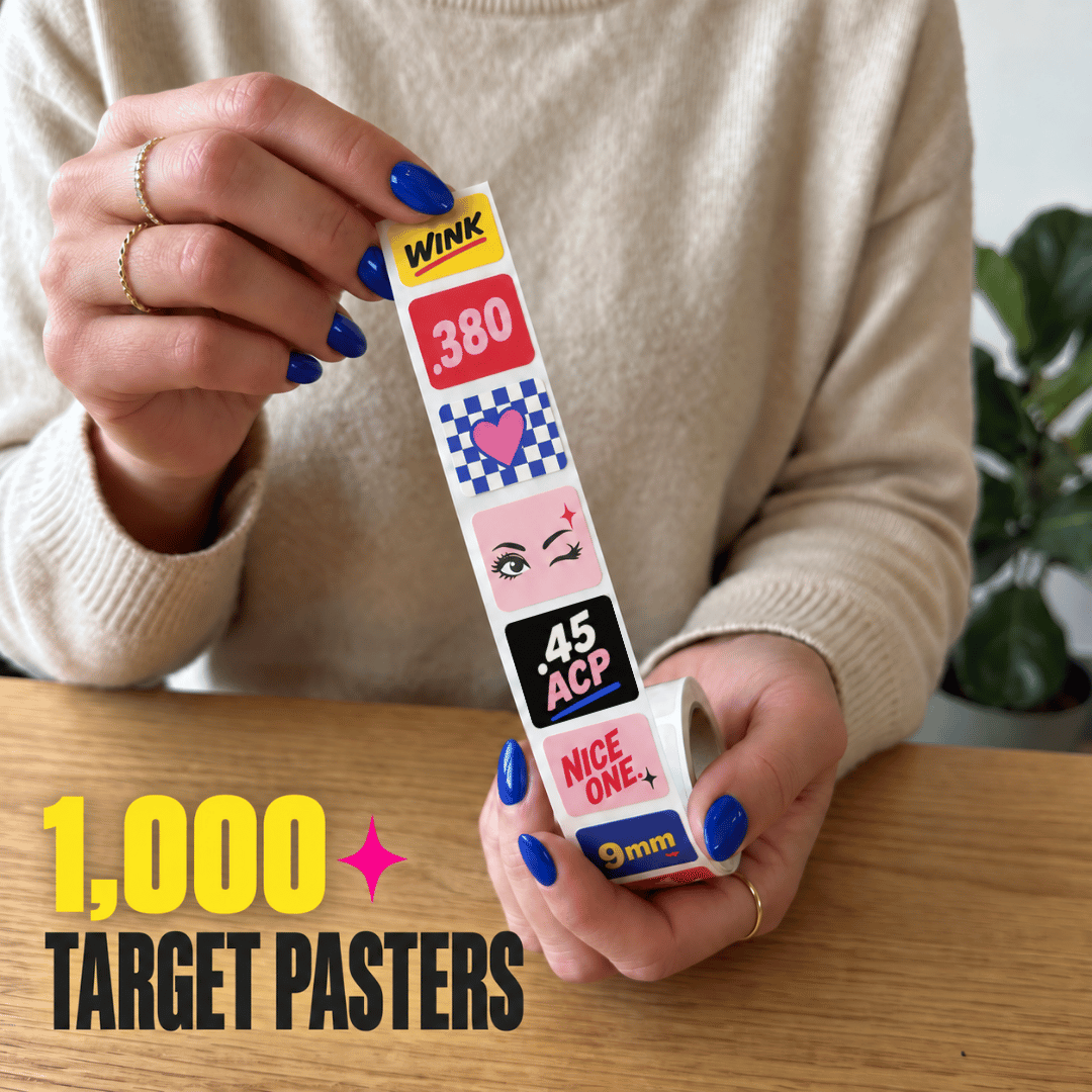 Woman holding a roll of 1,000 STRAPPED HELL YES target pasters with colorful designs including WINK, NICE ONE, and 9mm.