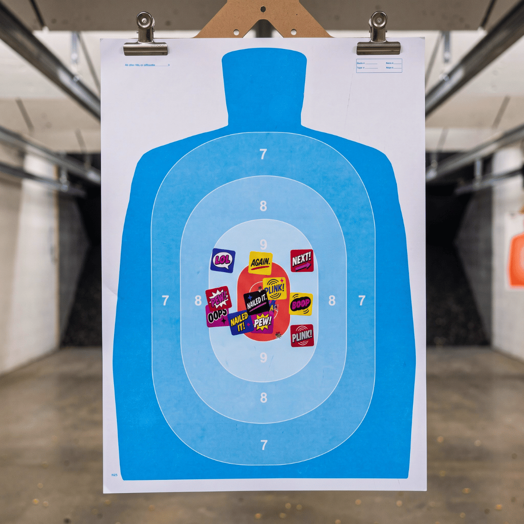 Blue silhouette shooting target at an indoor range covered with colorful STRAPPED PEWS &amp; PALS stickers over bullet holes near the center.