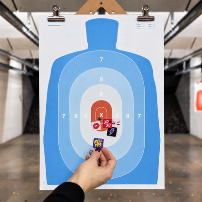 Hand placing a HELL YES sticker on a blue silhouette shooting target alongside colorful STRAPPED target pasters covering bullet holes.